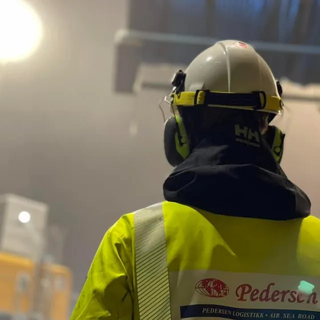 En arbeider i gul refleksvest og hjelm ser på løft av stor trommel innendørs.