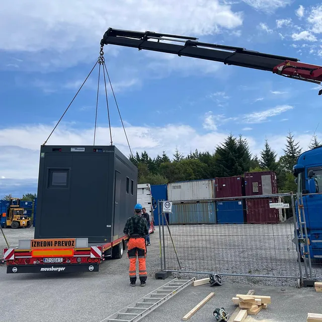 En mobilkran løfter en mørk modulbygning fra en tilhenger på et industriområde med containere i bakgrunnen.