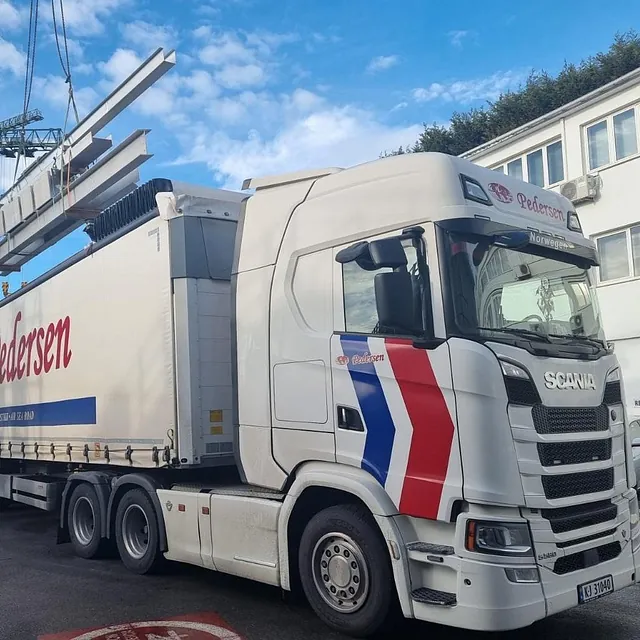 Lange ståldrager heises med kran over en Pedersen-lastebil utenfor industribygg.
